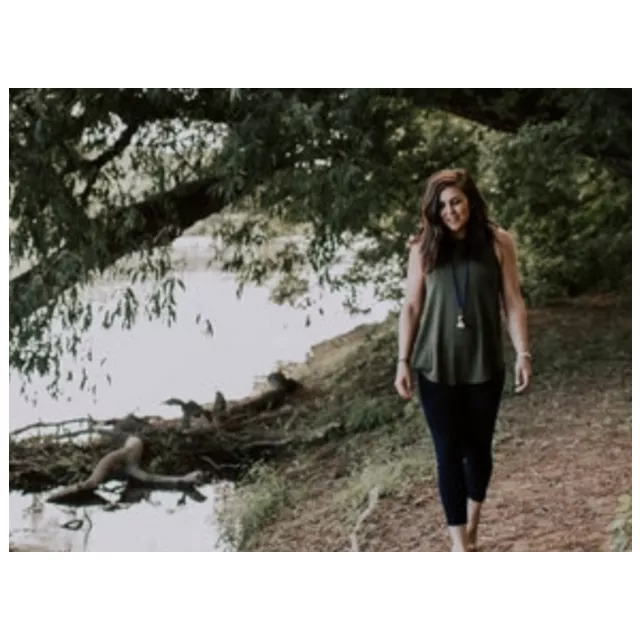 A brown hair female stands next to a river.  She is looking down.  Green trees are behind her. 